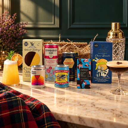 The Wee Cocktail Hamper with cocktail cans, biscuits and chocolate beside two served cocktails and a gold shaker.