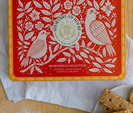 A red and orange Shortbread Selection tin by Shortbread House is shown on a wooden table with flour, a spoon, and cookies on a plate. The tin features white floral and bird illustrations with the brand logo in the center.