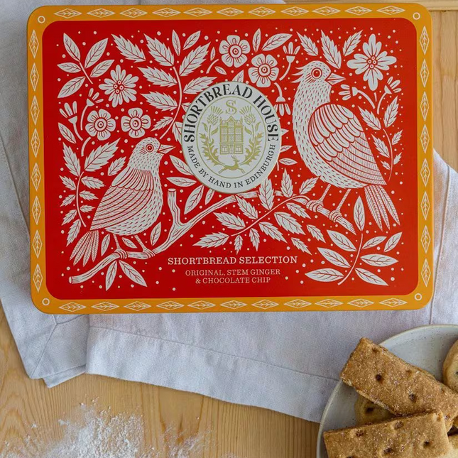 A red and orange Shortbread Selection tin by Shortbread House is shown on a wooden table with flour, a spoon, and cookies on a plate. The tin features white floral and bird illustrations with the brand logo in the center.