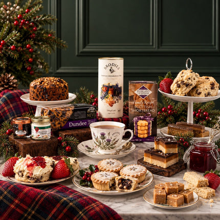 A festive Christmas afternoon tea spread on a marble table, featuring Sodi tea, Dundee shortbread, scones, mince pies, strawberries, jam, and a cup of tea, styled with green pine branches and a red plaid blanket against a dark green paneled wall.