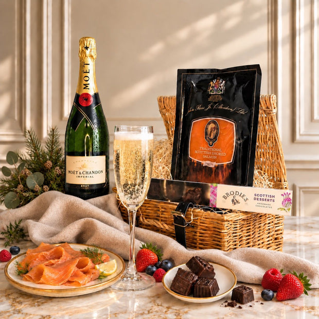 A luxury gift hamper set on a marble table featuring a bottle of Moët & Chandon Non Volontaire, a filled champagne flute, a wicker basket with a black bag of Brodie's Collection Ltd. chocolates, and plates of smoked salmon and chocolate desserts, styled with a beige cloth and greenery.