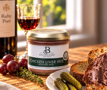 A jar of Ballancourt Salmon Terrine with dill sits on a wooden table beside a white plate of salmon terrine on toast, garnished with dill and pickled red onion, with a blurred kitchen background.