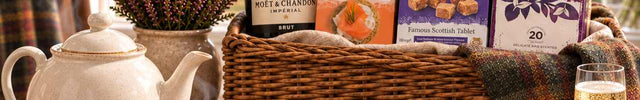A large woven basket gift hamper filled with assorted food items, including a bottle of Moët & Chandon, a jar of Marmalade, and boxes of biscuits, set on a wooden table with tea, bread, cheese, and a glass of sparkling wine in front of a window, Create Your Own Hamper