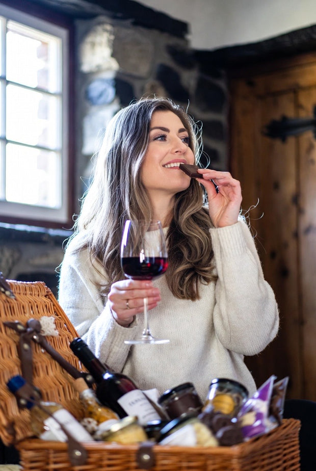 Woman enjoying her gift basket with a glass of red wine and a chocolate bar in a cozy indoor setting.