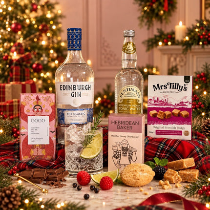 A festive holiday gift set arranged on a table in front of a decorated Christmas tree. The set includes a bottle of Edinburgh Gin, a bottle of Centimal Tonic Water, a box of Mrs.