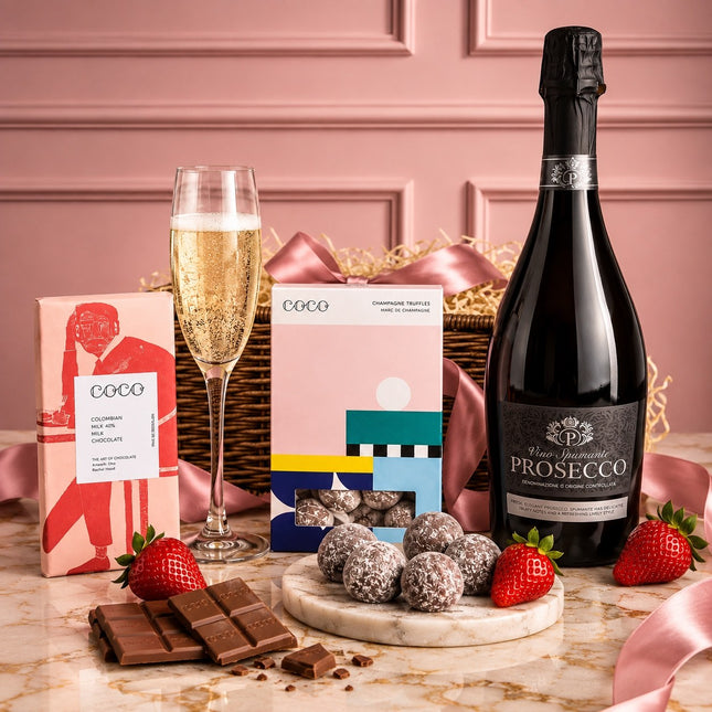 A luxury champagne and chocolate gift hamper featuring a bottle of Prosecco, a glass of bubbly champagne, boxes of Coco chocolates, chocolate bars, strawberries, and truffles arranged on a marble surface in front of a pink basket.