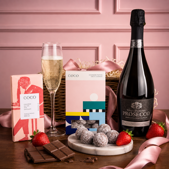Bottle of Prosecco, glass of champagne, chocolate bars, strawberries, and gift boxes on a wooden table with a pink background.