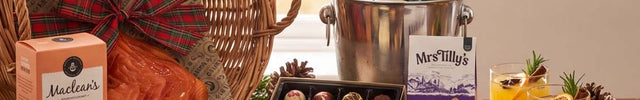 A gourmet gift hamper arranged on a rustic wooden table in front of a window. The set includes a large wicker basket with a red plaid bow, a box of Muslin's cheese, blue cheese, sliced carrots, chocolate truffles, a bottle of champagne in an ice bucket, two glasses of orange drink, and handwritten cards reading 'For Someone Special'.
