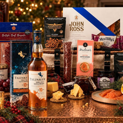 A Christmas-themed gourmet gift hamper arranged on a copper table in a warmly lit living room. The set includes a bottle of Talisker 10 Year Single Malt Scotch, John Ross & Son whisky, British Beef Brisket, Hawshead Glaid, MacTilly's Irish Soda Pies, Murrin's Baked Goods, and other seasonal treats, surrounded by candles, ornaments, and a plaid runner.