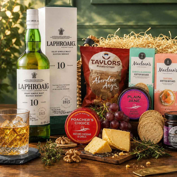 A styled gift basket arrangement on a wooden surface featuring a Laphroaig 10 Year single malt whisky bottle, its box, a glass of amber whisky, and assorted food items including Taylor's potato chips, Maclean's snacks, Poacher's Choice cheese, grapes, crackers, walnuts, and rosemary, set against a blurred green background.
